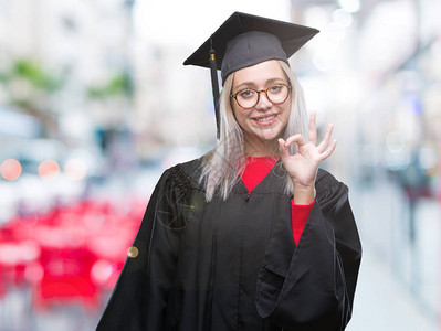 身着毕业制服的金发年轻女子在孤立的背景中微笑地用手和指做一个好标记图片