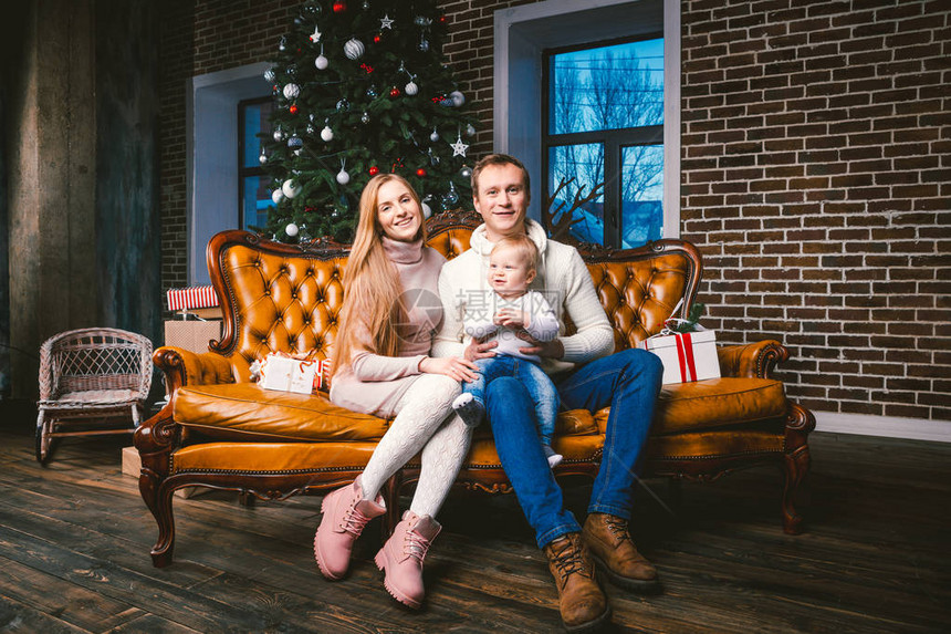 Mood庆祝高加索年轻妈爸和儿子一岁时坐在圣诞树附近的客厅里家的棕色皮沙发上图片