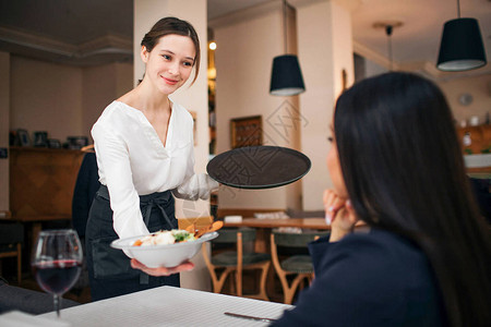 令人愉快的年轻女服务员与坐着的顾客站在餐桌旁图片