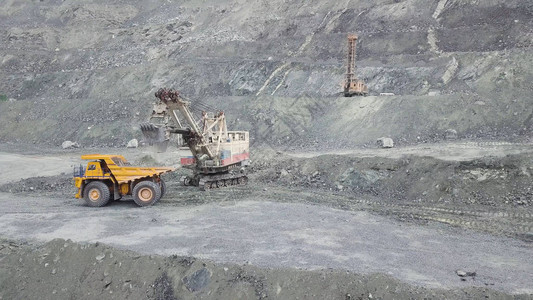 在灰色采石场采矿业装载石矿时的挖掘机和自卸车重型采矿挖掘机将岩石矿装入翻斗式大型采矿卡车背景
