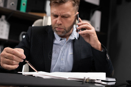 身穿黑色夹克和浅色衬衫的可敬男子在电话中交谈并在文件中做笔记商人在办公室的画像现代世界的经营理背景图片