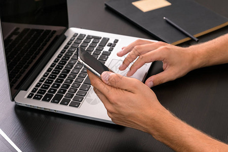 男人在用旧的木制桌子使用笔记本电脑时手打智能电话手打键盘顶级视图图片
