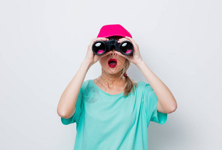 穿着粉红色帽子和蓝色T恤的年轻美女寻找白色背景上戴望远镜的东图片