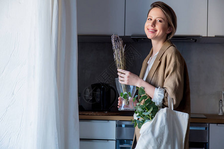 美丽的女孩在夹克与手提袋和植物在家里的厨房里生活方式的健图片