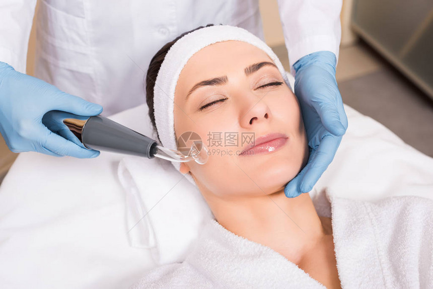 美容师在美容院用设备对脸颊上的女人做darsonvalization图片