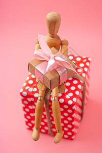 木制玩具模型拿着礼物坐在粉红色背景的红色圆点盒子上情人节和妇女节的图片