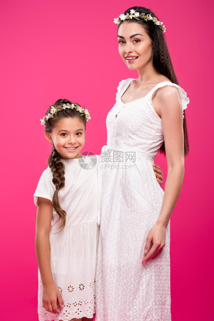 穿着白裙子和花圈的美丽的幸福母亲和女儿在粉红色上被隔绝的相机图片