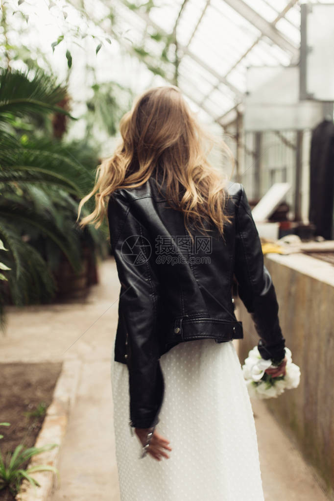 身穿黑色皮夹克和白裙子的年轻美女在旧温室走来去时手握着图片