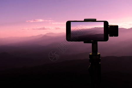 三脚架上使用智能手机旅行者在山地自然背景日落时拍摄照片图片