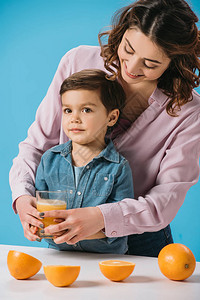 快乐的母亲给全杯新鲜橙汁给被蓝色隔离的可爱小儿子图片