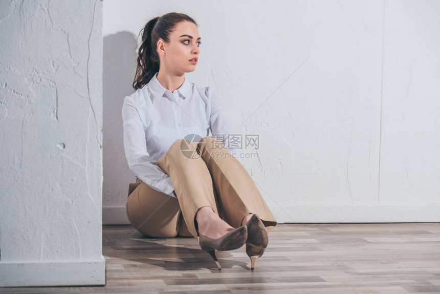 穿着白上衣和米色短裤的不开心妇女坐在家中白色墙附近的地板上图片