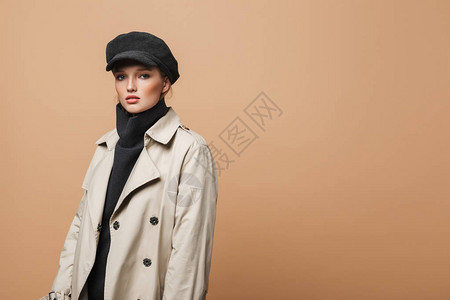 穿着经典海沟大衣和帽子的年轻美女仔细地在米色背景图片