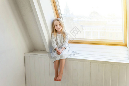 穿着白色连衣裙的可爱甜美微笑小女孩坐在家里明亮客厅的窗台上图片
