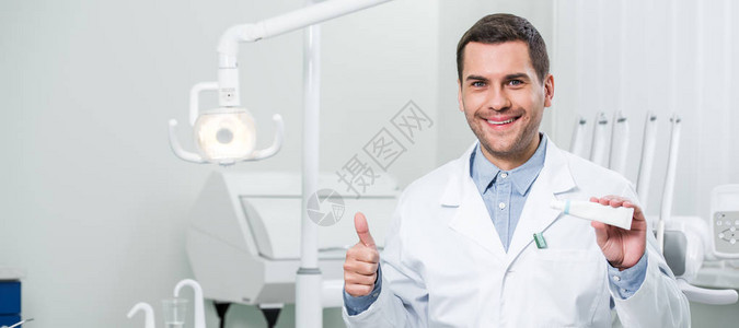 英俊的牙医在牙科诊所内拿着牙膏和大图片