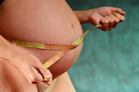 蓝色背景上穿着粉色内裤的怀孕女孩在厘米磁带上测量腹部的体积图片