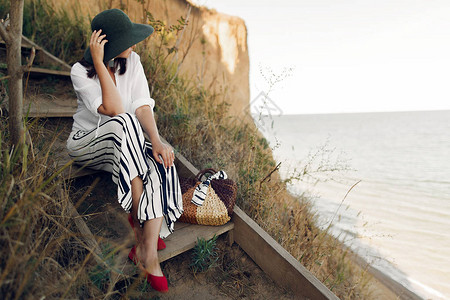 时尚的女孩坐在阳光明媚的海上滩悬崖上的木制楼梯上图片