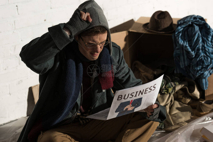 无家可归的男子在被垃圾围着坐时阅读商业报图片