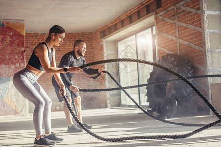 交替推肩年轻的健身夫妇在服做量来找到心血管锻炼使用训练绳在健身房背景