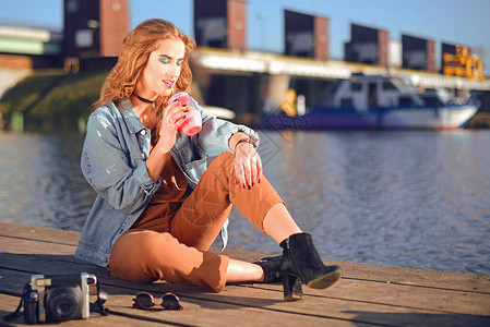 美丽的女孩坐在河边的码头上时髦模型在短项链与cerly头发女孩在炎热的晴天喝柠檬水夏日风情有老照相机的女孩背景图片