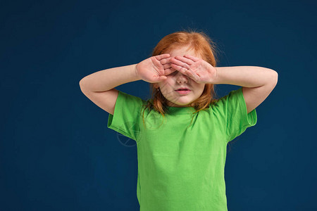 小红发女孩从明光或阳光蓝背景绿色T恤上遮着眼图片