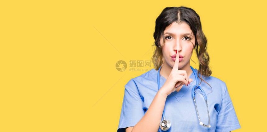 身穿医疗制服的年轻成年女医生要求用手指嘴唇保持安静沉默图片
