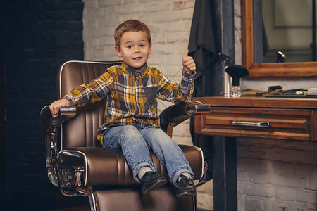一个穿着衬衫和牛仔裤坐在椅子上与理发师工作场所对面的时髦小男孩的肖像图片