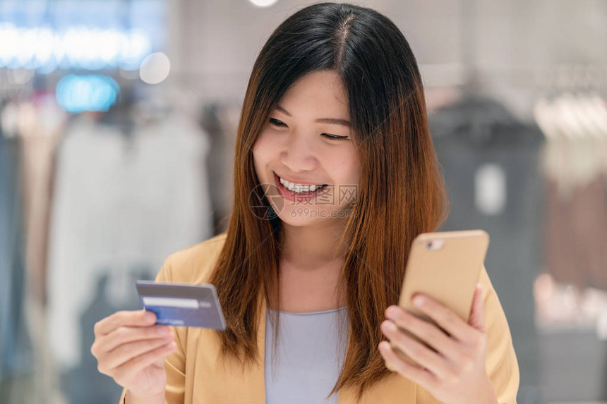 亚洲女使用带手机的信用卡在服装店背景下的百货公司网上购物技术货币钱包和在线支付概念图片
