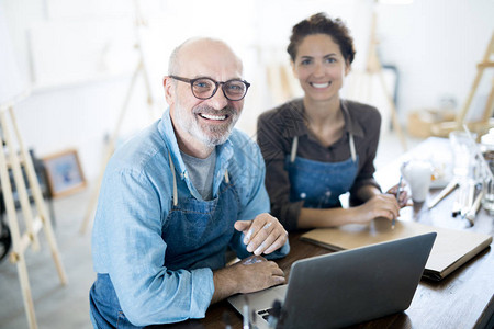 微笑的老人和他穿着工作服的同事坐在工作室或工作室的笔记本电图片