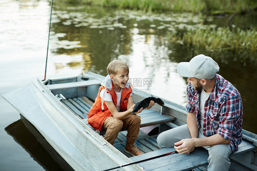 快乐的男孩给他父亲看大鱼他坐着在船上聊天图片