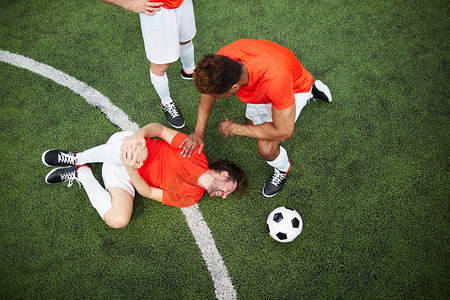 两个球友站在受伤的足球运动员旁边躺在绿色图片