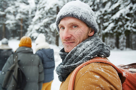 穿着雪帽和围巾的帅年轻男子徒步旅行者在看冬季森林的相机时转过身去图片