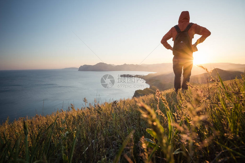 年轻的常设男子背着包在五颜六色的日落天空海边的石头上的徒步旅行者美丽的风景与运动型男子在日落时岩石海和云运图片