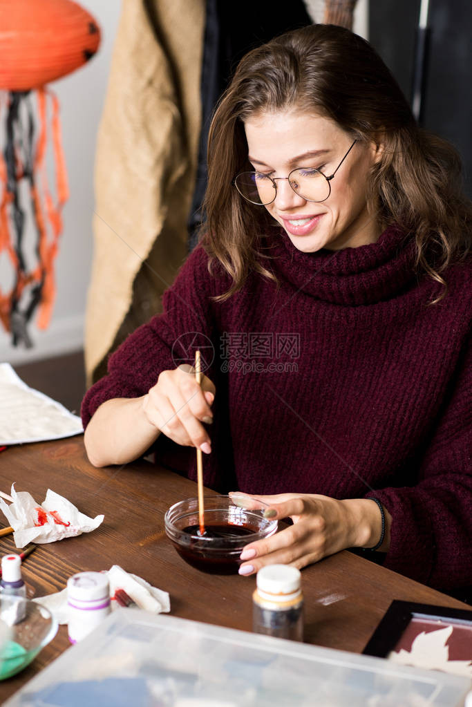 快乐兴奋的创意年轻女设计师戴着眼镜坐在桌边混合油漆为万圣节装饰制作血色图片