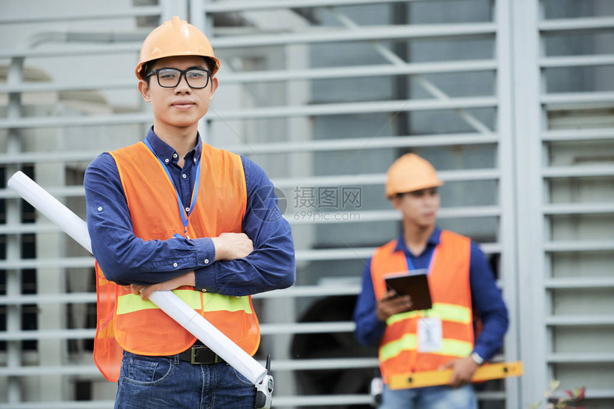 身着头盔和腰衣的有吸引力的亚洲男子在同事附近建筑工地站立时图片