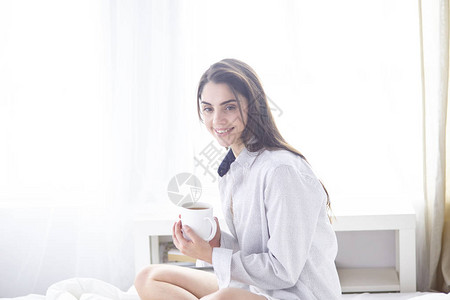 穿着黑丝袍睡在床上喝早间咖啡白杯子妇女节日假期的年轻美丽的图片