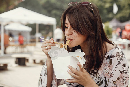 时尚的时髦女孩用竹筷子从纸箱里吃蔬菜炒锅面条亚洲街头美食节快乐的波西米亚女人在外卖背景图片