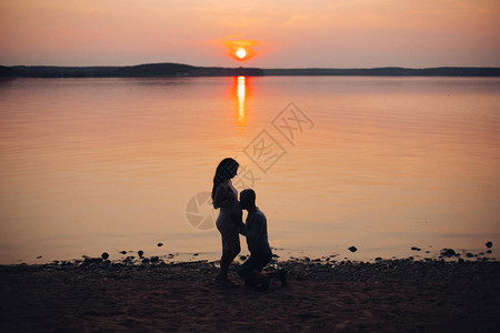 孕妇和她的丈夫在海边日落时的剪影男人和亲吻她的肚子期待一个婴儿的概念图片