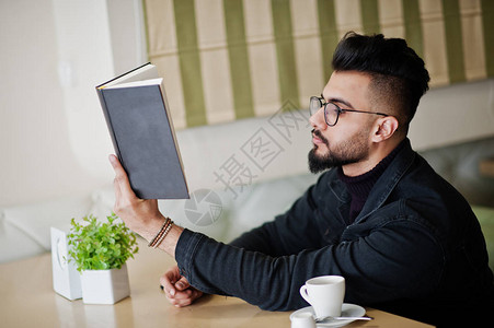 阿拉伯男子穿着黑色牛仔裤夹克和眼镜坐在咖啡馆里看书喝咖啡时尚图片