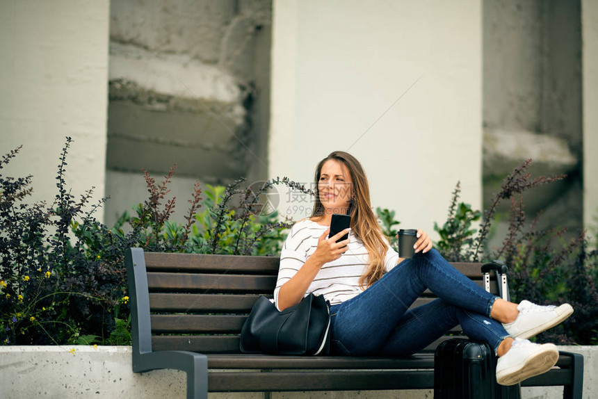 坐在长椅上在车站等候时用智能电话的女士图片