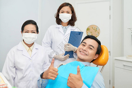 亚裔快乐的男子展示拇指在牙科椅上和亚洲女穿着医疗图片