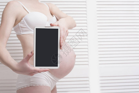 穿着内衣的孕妇持有并展示数字平板电脑图片