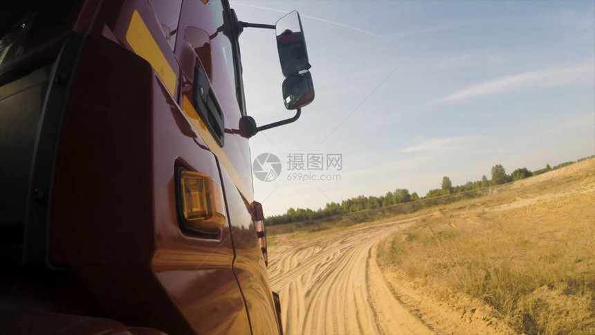 红色卡车驾驶室在尘土飞扬的未铺砌乡村道路上沿绿树前进的侧视图图片