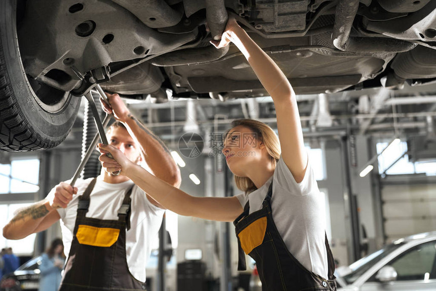站在汽车下和在车间工作的专业机械师年轻女工与强壮的男同事在汽车服务中检查汽车底盘维护图片