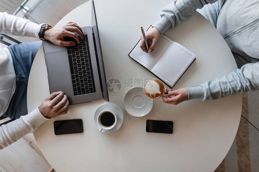 在男人和女人的商务会议上工作流程的顶部视图白色桌子上的顶视图与笔记本电脑与两杯咖啡和手机的记事本在非正图片