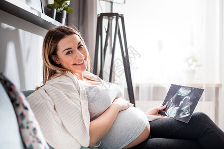 坐在沙发上的孕妇用超声波图像看着她的婴儿图片