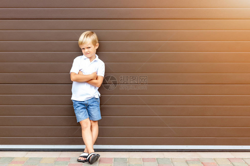 穿着休闲服的可爱脾气暴躁的金发孩子站在棕色车库门旁愤怒的男孩在房子附近双臂交叉尴尬的年龄和育儿理念孩子图片