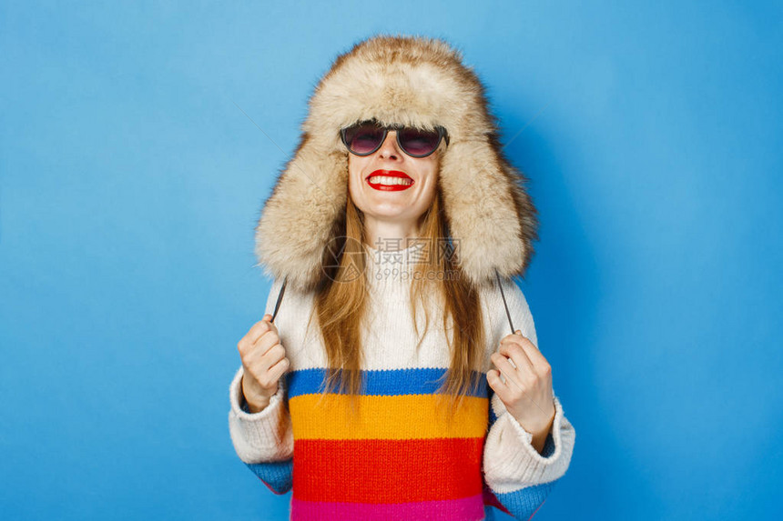 一个戴着毛皮帽眼镜和蓝色背景毛衣的快乐脸的女孩冬季寒假图片
