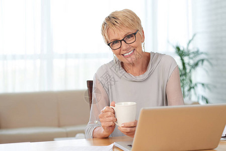 微笑的年长妇女在家里喝咖啡和做笔记本电图片