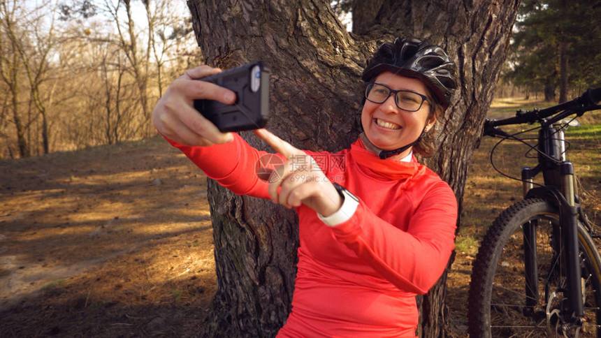 年轻的高加索女运动员旅游自行车手使用智能手机自拍坐在城外针叶林的树旁女运动员用她的山图片