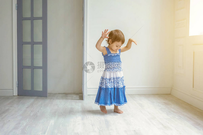 穿着蓝色连衣裙的可爱甜美微笑小女孩在家里明亮的客厅里跳舞图片